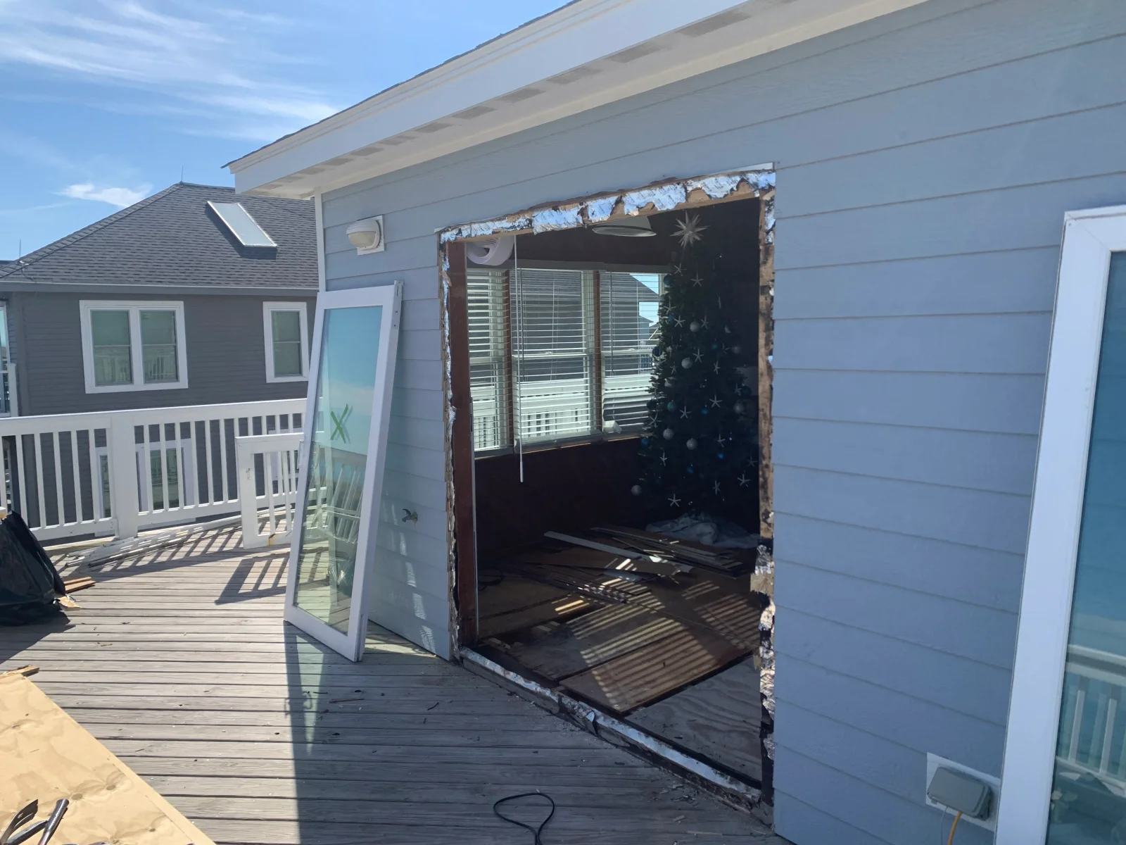 Exterior wall opened for door replacement, blue fiber cement siding, exposed framing, white framed glass door panel, weathered deck