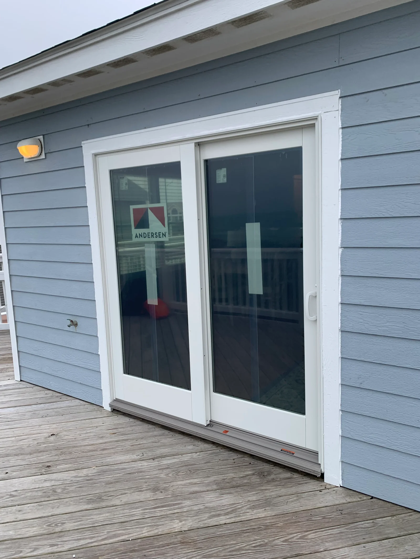 White-framed Andersen two-panel sliding patio door with clear glass in blue lap siding, opening onto a wood deck